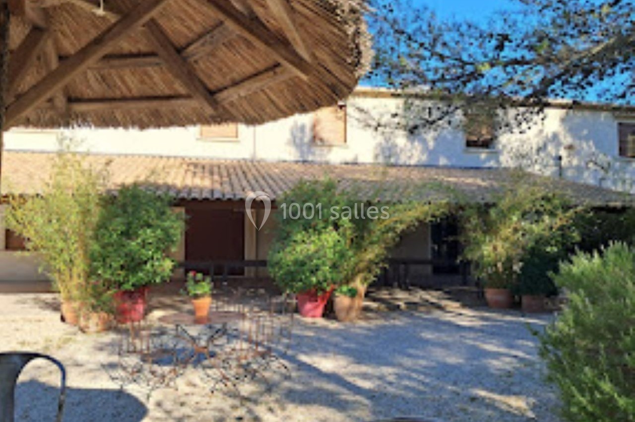 Cour extérieure ensoleillée avec des plantes en pots, des chaises en métal et un bâtiment à toit de tuiles en arrière-plan.