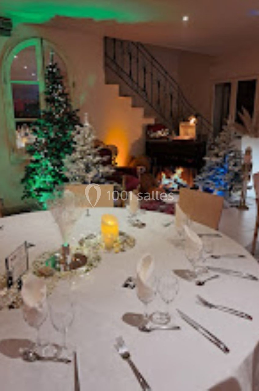 Salle décorée pour une réception avec table dressée, sapins illuminés et escalier en arrière-plan.