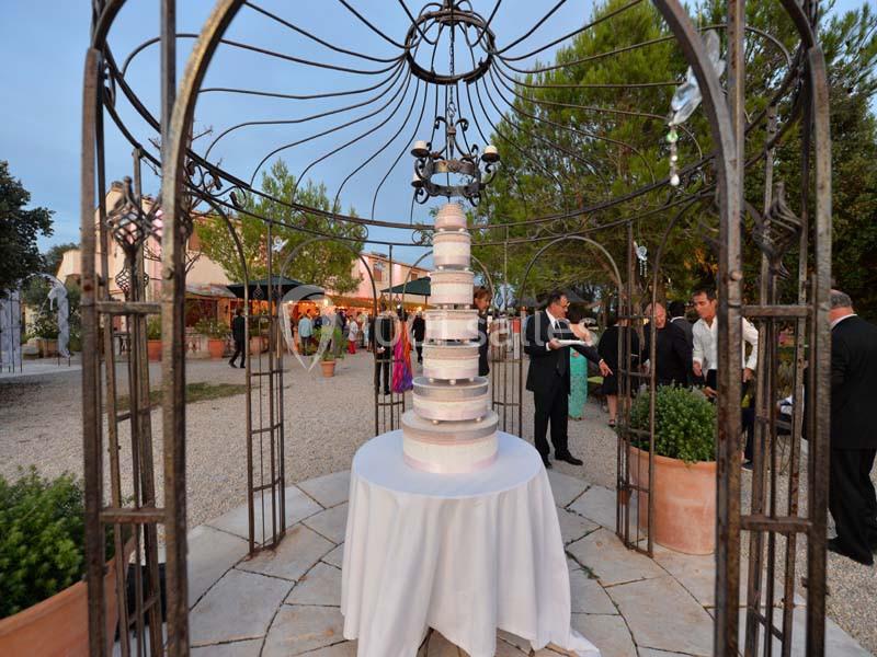 Gâteau de mariage à plusieurs étages sous une structure métallique, entouré d'invités dans un jardin en soirée.