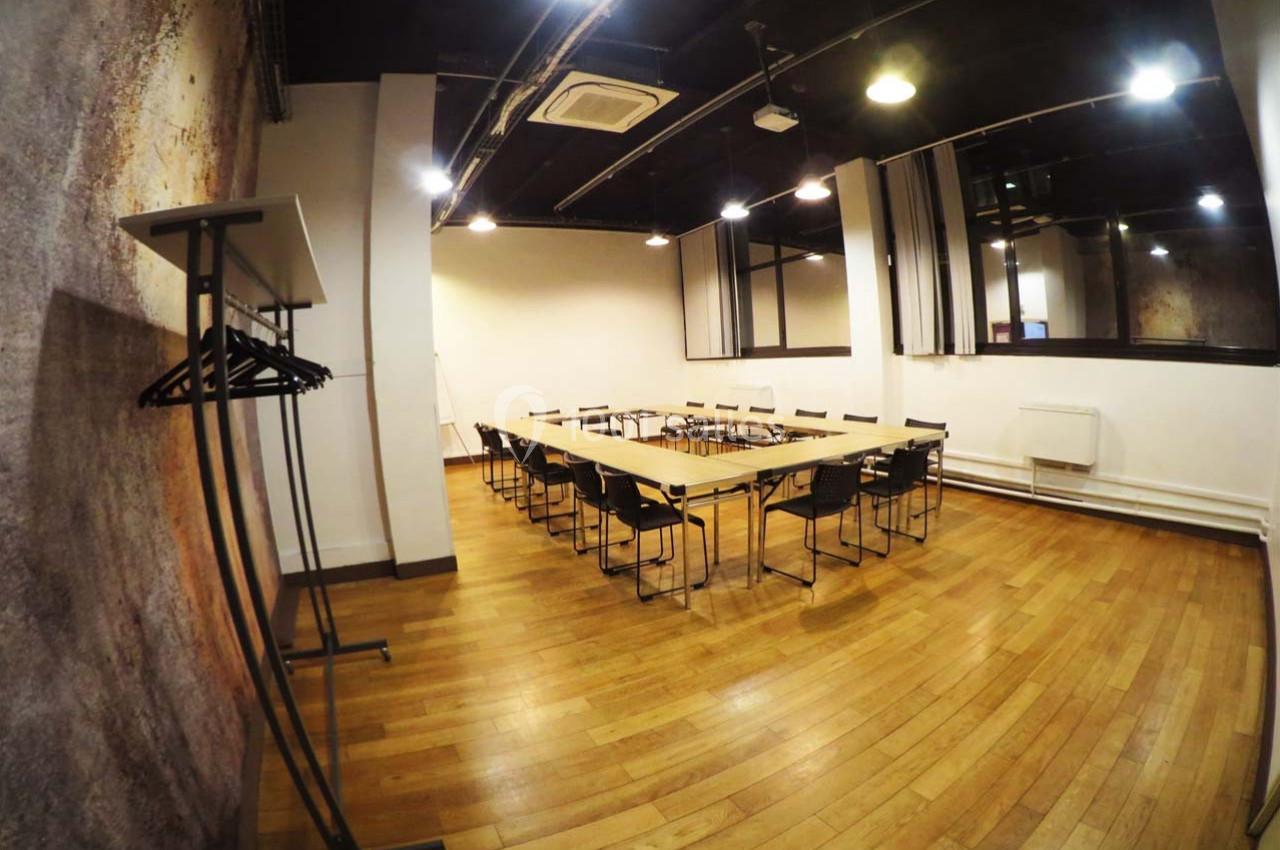 Salle de réunion avec tables disposées en rectangle, chaises noires, parquet en bois et éclairage au plafond.