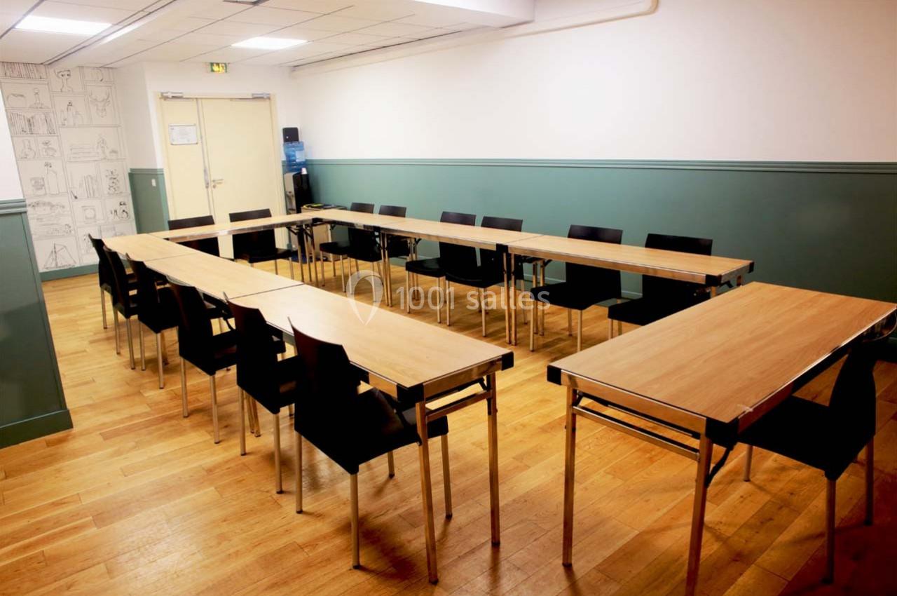 Salle de réunion avec tables en bois disposées en U, chaises noires et murs verts dans un espace lumineux.