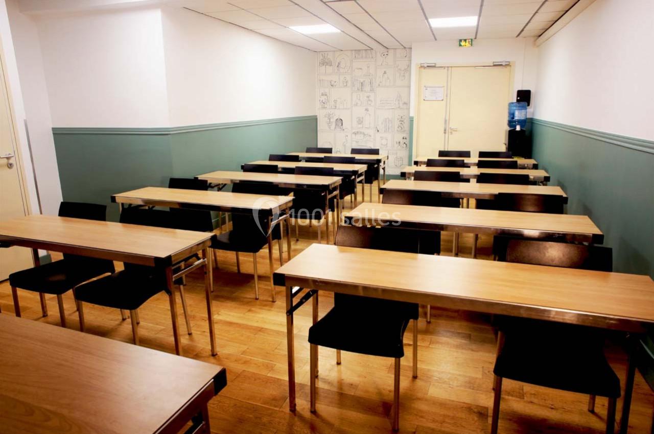 Salle de classe vide avec des tables en bois, des chaises noires et un mur décoré de dessins au fond.
