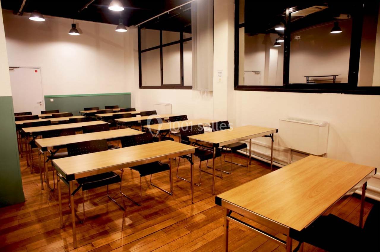 Salle de classe vide avec des tables en bois, des chaises noires et un éclairage suspendu.