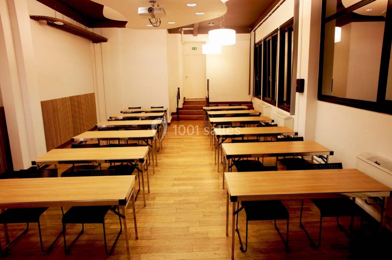 Salle de formation avec des tables en bois alignées, des chaises noires et un éclairage chaleureux.