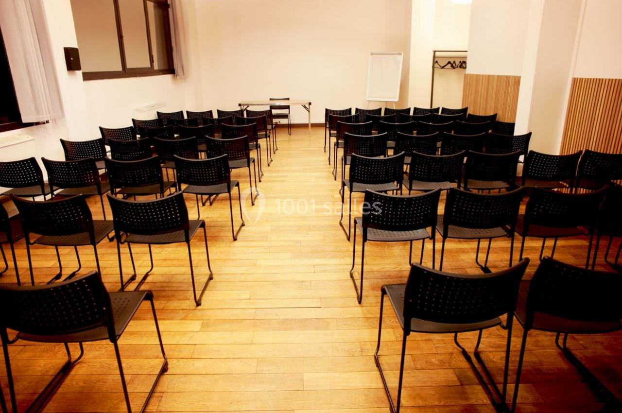 Salle vide avec des rangées de chaises noires alignées sur un sol en bois, tableau blanc en arrière-plan.