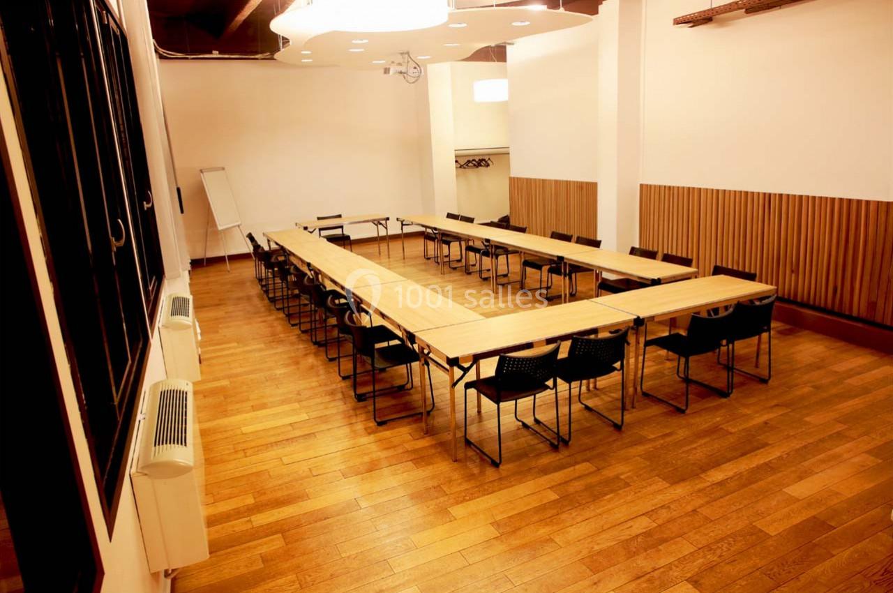 Salle de réunion lumineuse avec tables disposées en U, chaises noires, parquet en bois et murs clairs.