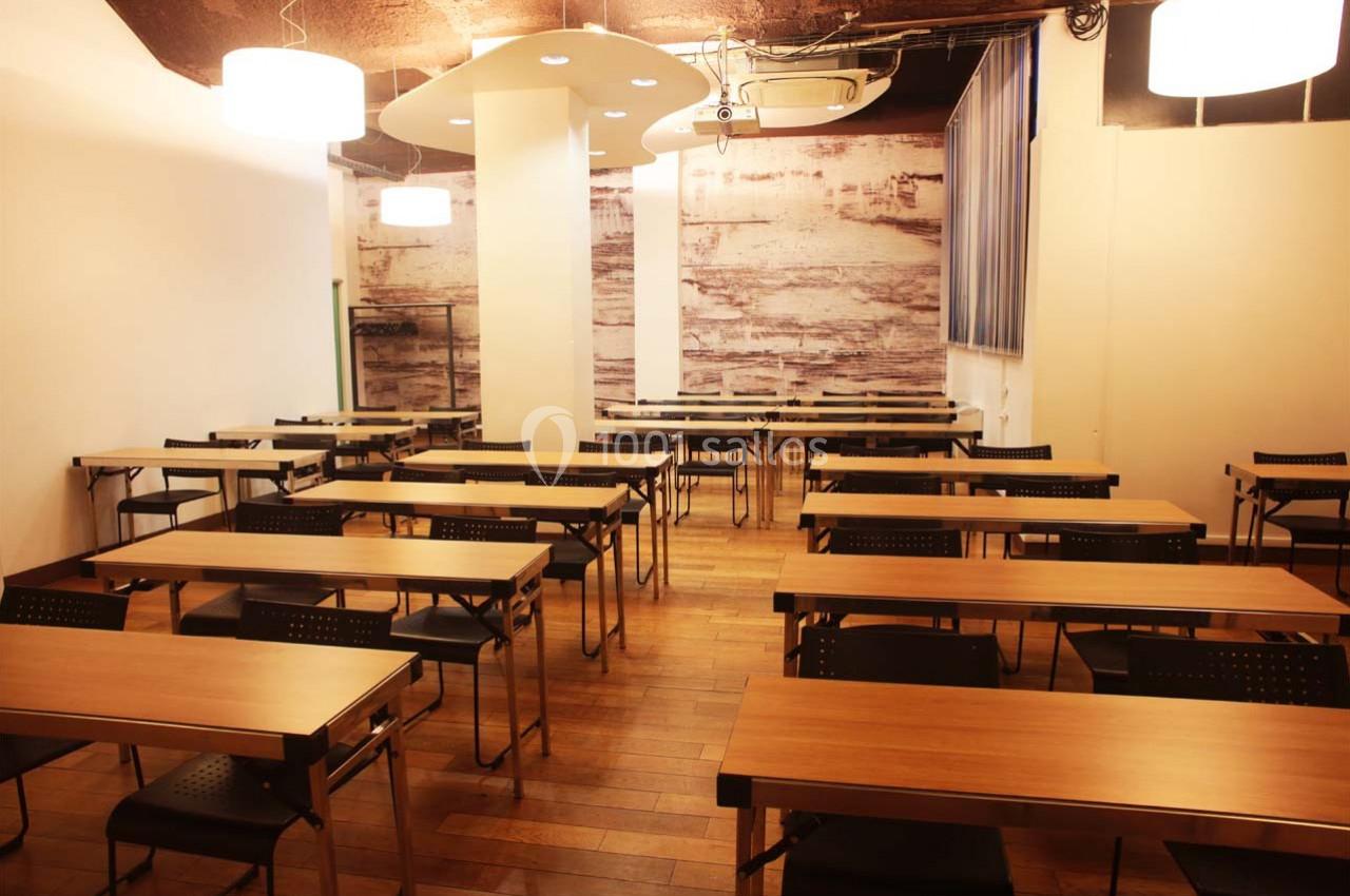 Salle de classe vide avec des tables en bois, des chaises noires et un éclairage chaleureux.