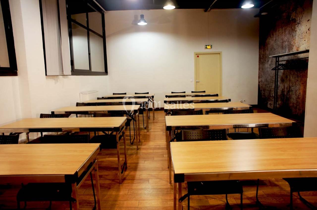 Salle de classe vide avec des tables en bois, des chaises noires et un éclairage suspendu.
