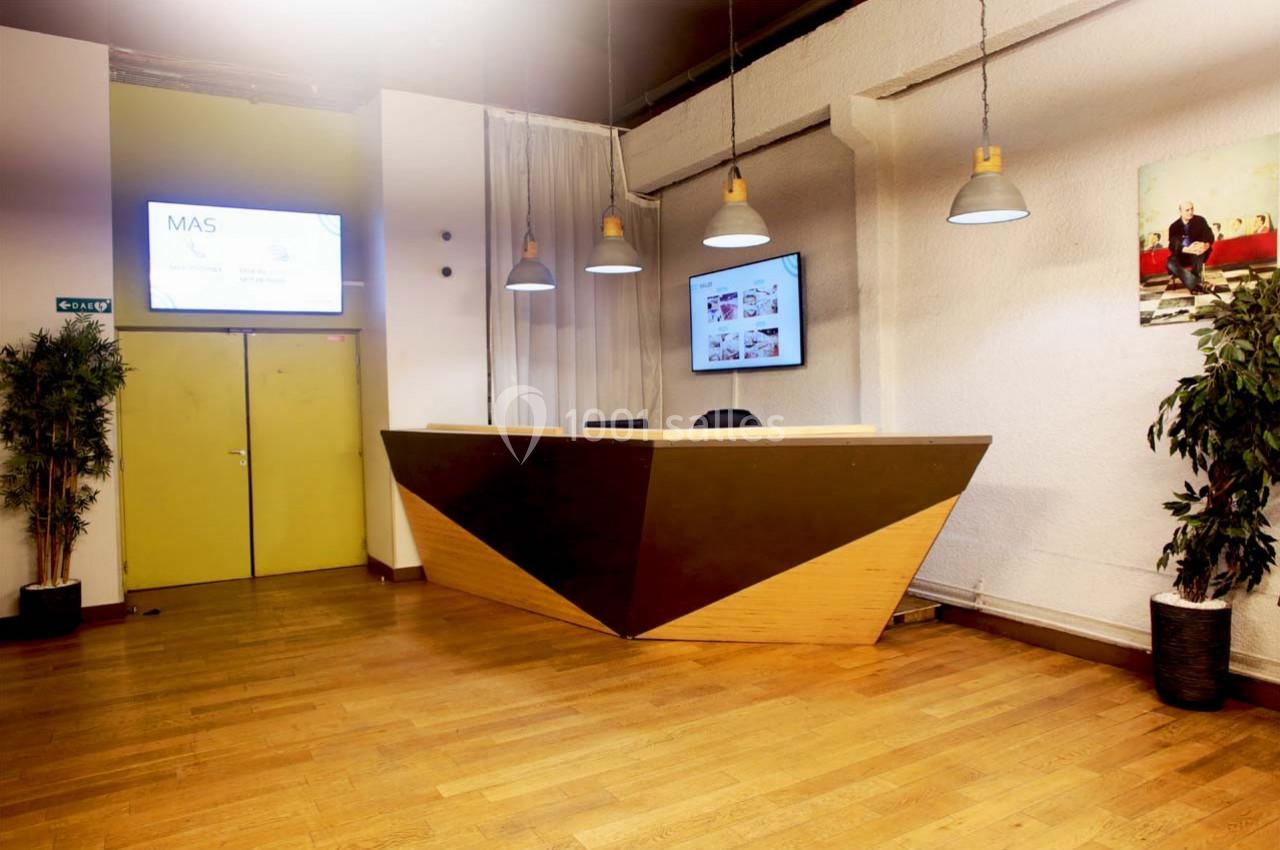 Bureau d'accueil moderne avec comptoir en bois, écrans muraux, plantes et éclairage suspendu dans un espace lumineux.