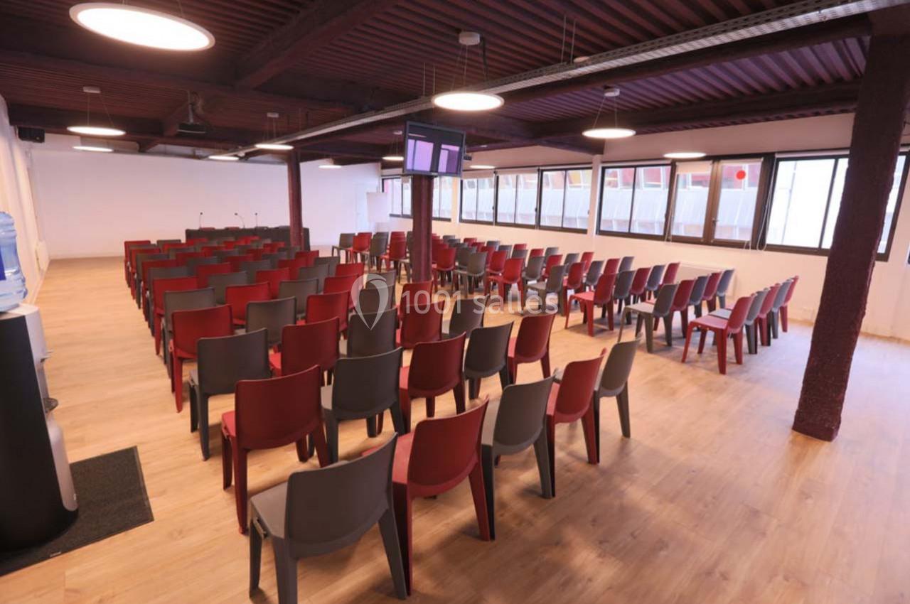 Salle de conférence avec des rangées de chaises rouges et grises, éclairée par des plafonniers et des fenêtres latérales.