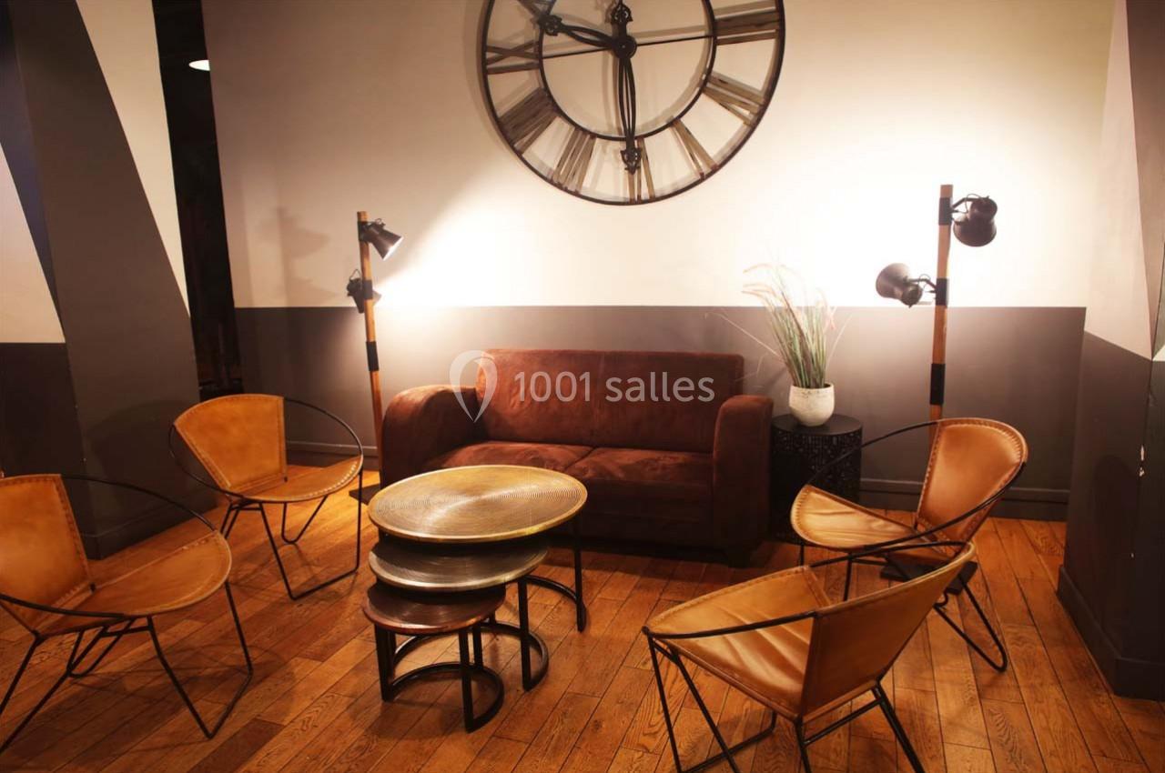 Espace salon avec canapé en velours marron, chaises en cuir, tables rondes et grande horloge murale.