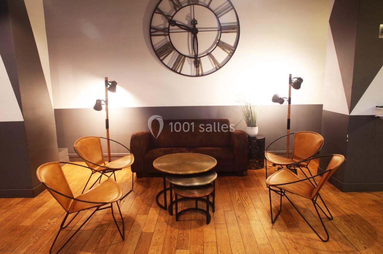 Salon avec canapé marron, quatre fauteuils en cuir, tables basses rondes et une grande horloge murale.