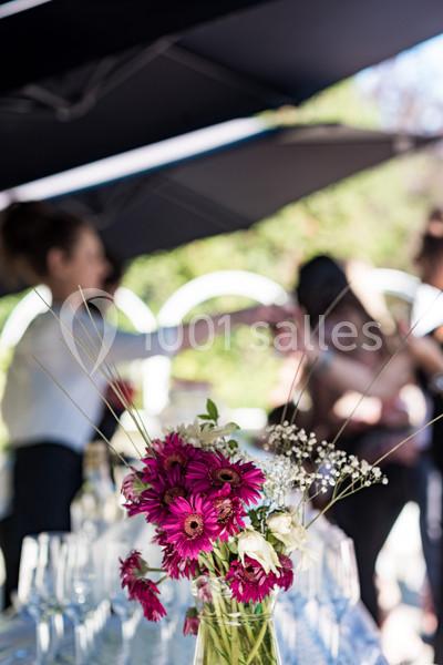 Vase de fleurs roses et blanches sur une table, avec des personnes floues en arrière-plan sous des parasols noirs.