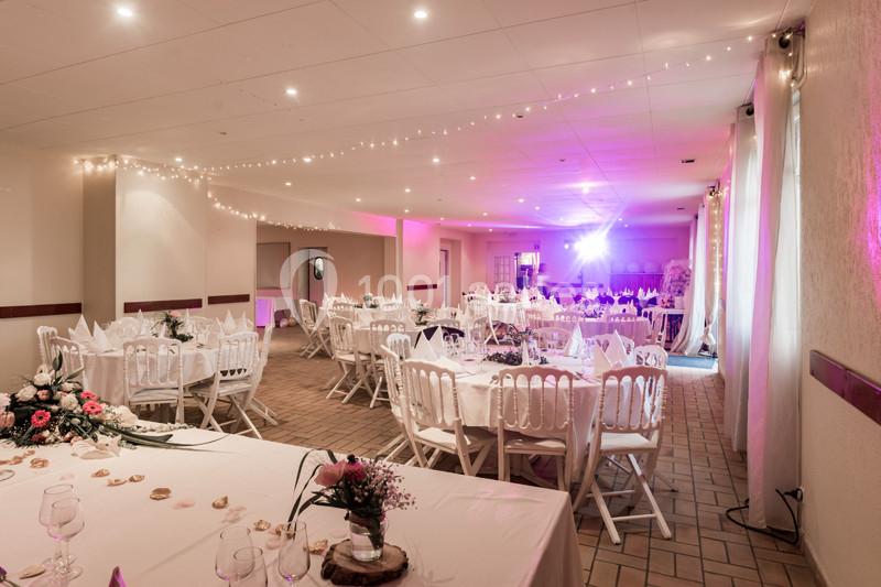Salle de réception décorée avec des tables rondes dressées, des chaises blanches et des guirlandes lumineuses au plafond.