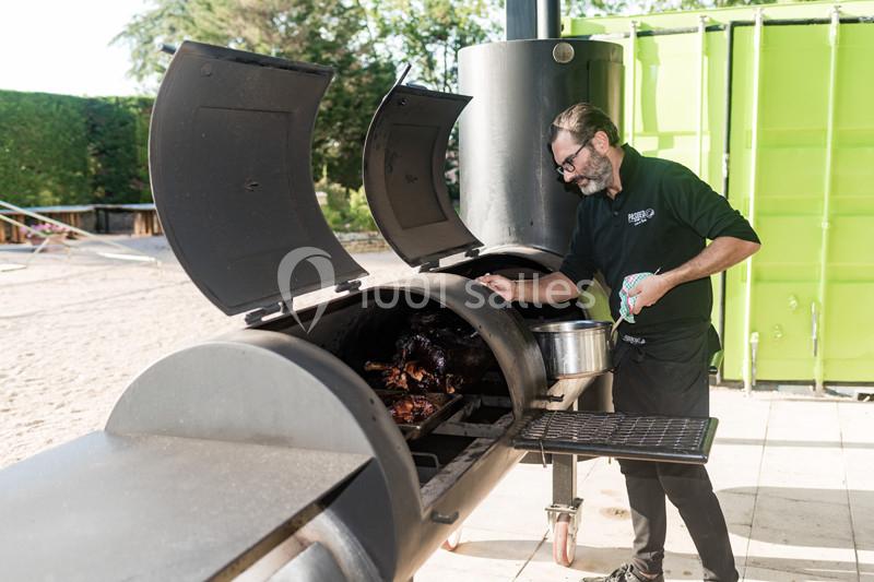 Un homme cuisine de la viande dans un grand fumoir métallique en extérieur, près d'un conteneur vert.