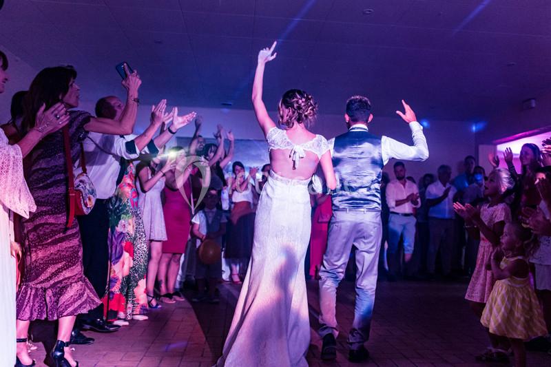 Un couple de mariés danse devant des invités applaudissant dans une salle éclairée par des lumières colorées.