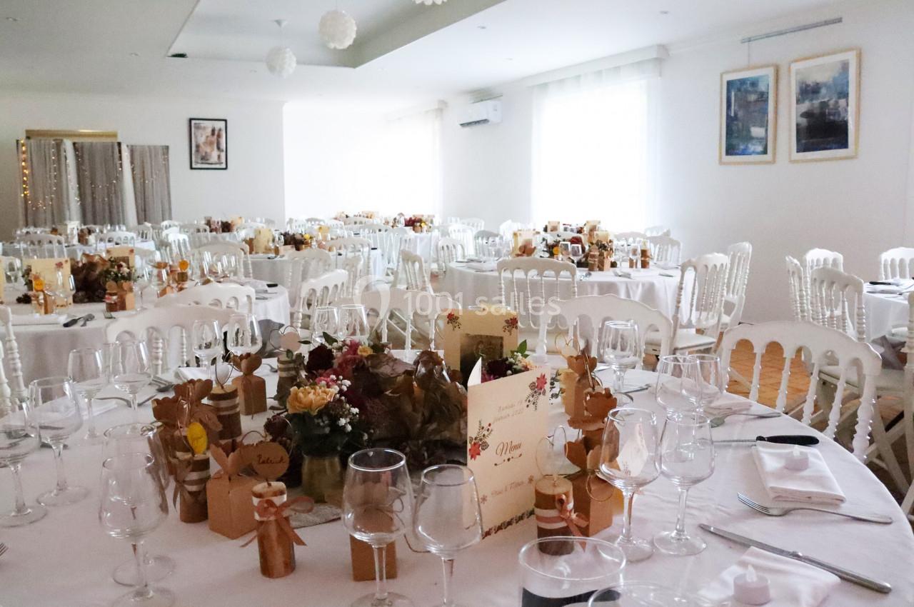 Salle de réception décorée pour un événement, avec tables dressées, chaises blanches et centres de table fleuris.