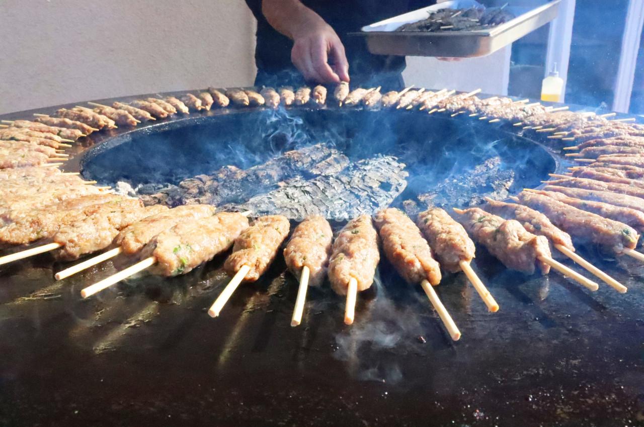 Brochettes de viande grillées sur un barbecue circulaire, avec une personne tenant un plateau en arrière-plan.