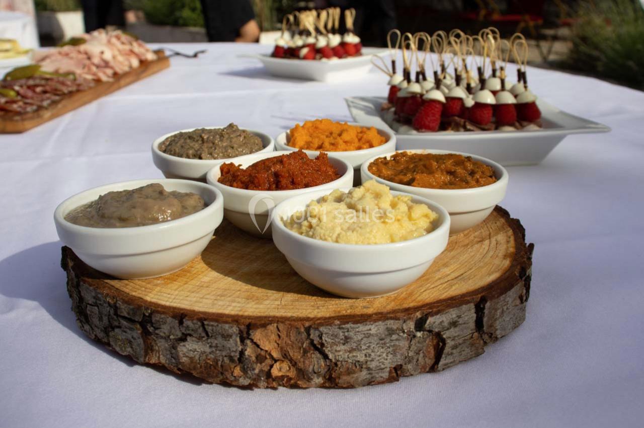 Assortiment de tartinades dans des bols blancs posés sur une rondelle de bois, avec des amuse-bouches en arrière-plan.