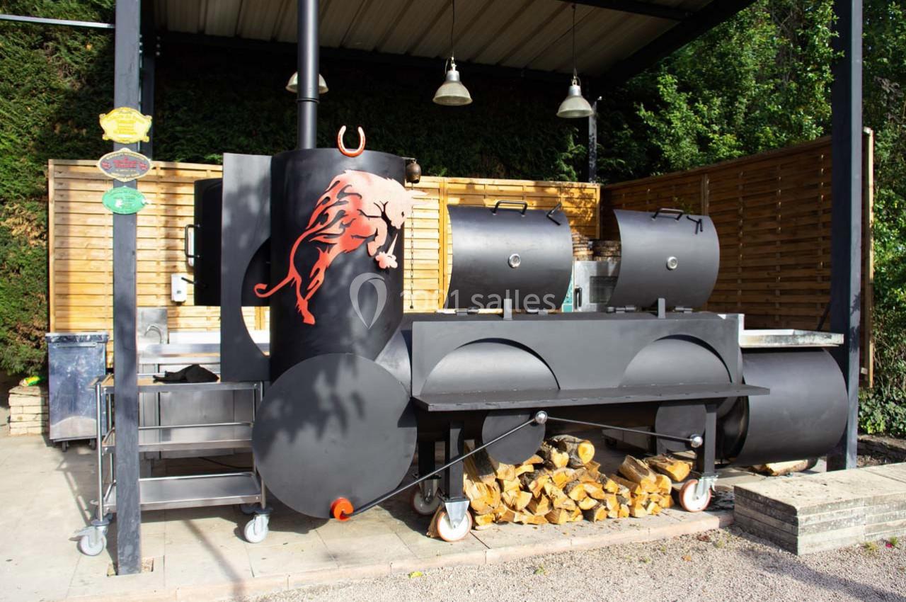 Barbecue fumoir noir en métal avec un motif de taureau rouge, abrité sous un auvent, entouré de bois de chauffage.
