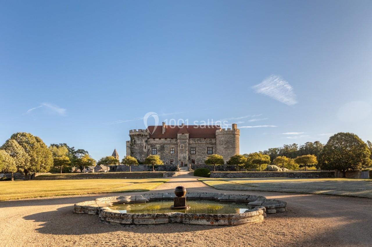 Vue d'un château en pierre avec un toit rouge, entouré d'arbres, et un bassin ornemental au premier plan.