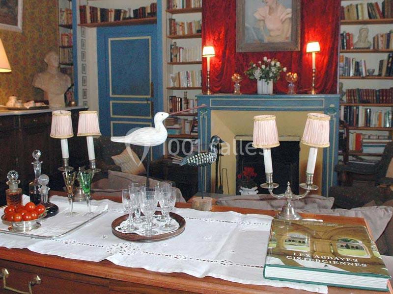 Intérieur d'un salon avec cheminée, bibliothèque, table décorée de chandeliers, carafes, verres et objets décoratifs.