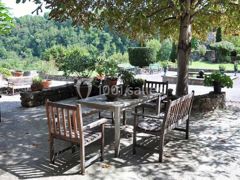Table et chaises en bois sous un arbre dans un jardin, entourées de plantes en pot et d'une vue sur la végétation.