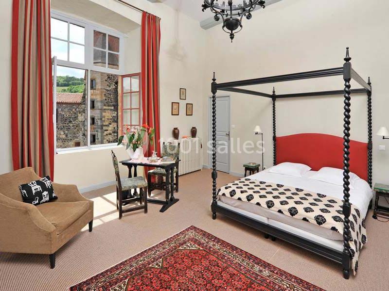 Chambre lumineuse avec lit à baldaquin, fauteuil, table dressée et grande fenêtre donnant sur un paysage verdoyant.