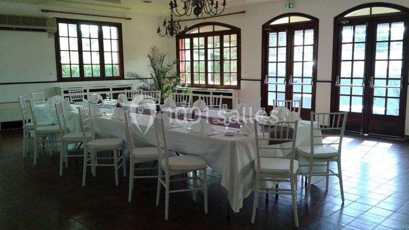 Salle lumineuse avec tables dressées en blanc, chaises blanches et grandes fenêtres donnant sur l'extérieur.