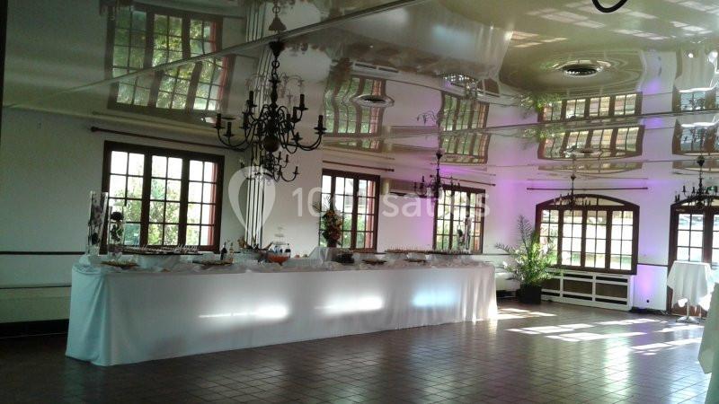 Salle lumineuse avec grandes fenêtres, plafond réfléchissant, lustres et une longue table dressée pour un buffet.