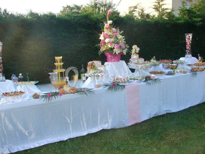 Table de buffet en extérieur avec nappes blanches, fontaine à jus, fleurs roses et divers plats disposés.
