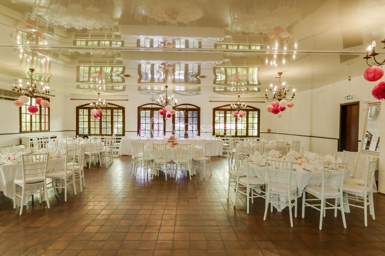 Salle de réception lumineuse décorée de lanternes roses, avec tables rondes dressées et chaises blanches.