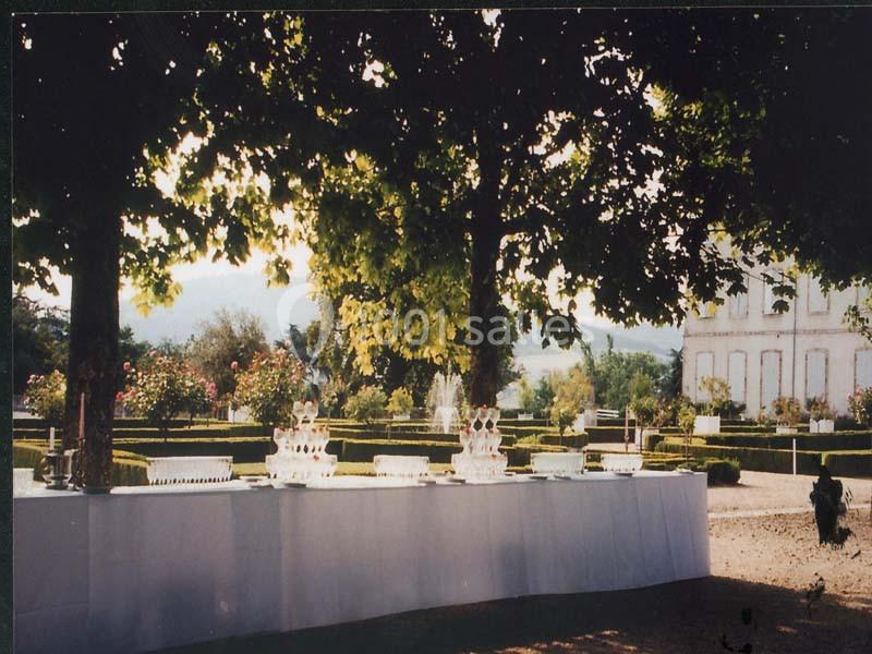 Table dressée en extérieur sous des arbres, avec des verres empilés et un jardin aménagé en arrière-plan.