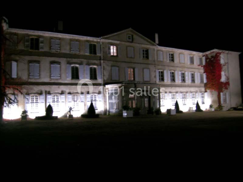 Façade d'un grand bâtiment ancien éclairé la nuit, avec des fenêtres et des plantes en pot devant.