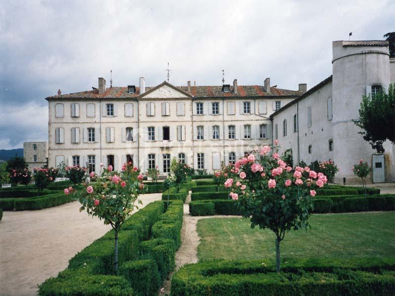 Façade d'un grand bâtiment ancien entouré de jardins symétriques avec des rosiers et des haies taillées.