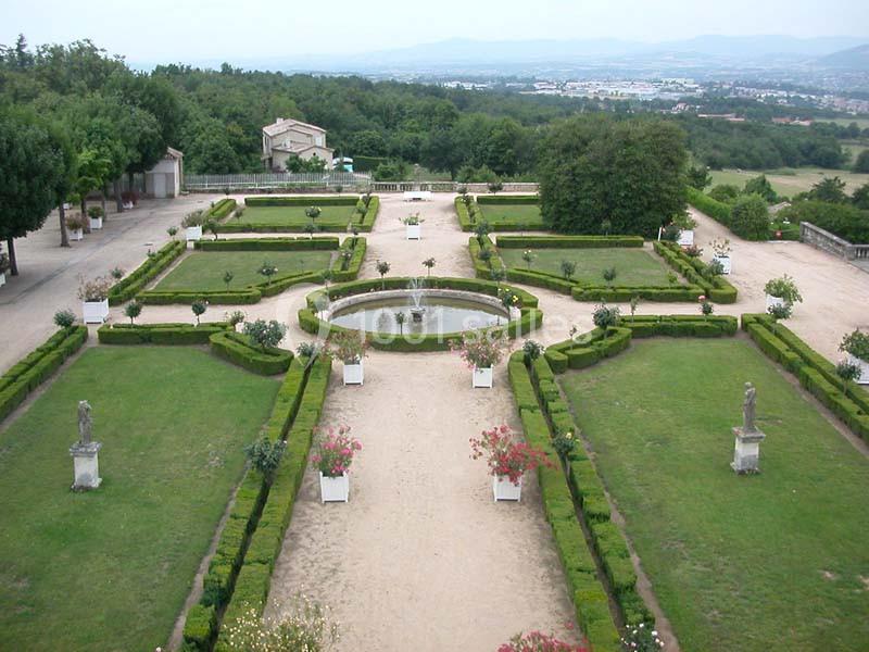 Vue d'un jardin symétrique avec allées, haies taillées, statues, bassin central et paysage vallonné en arrière-plan.