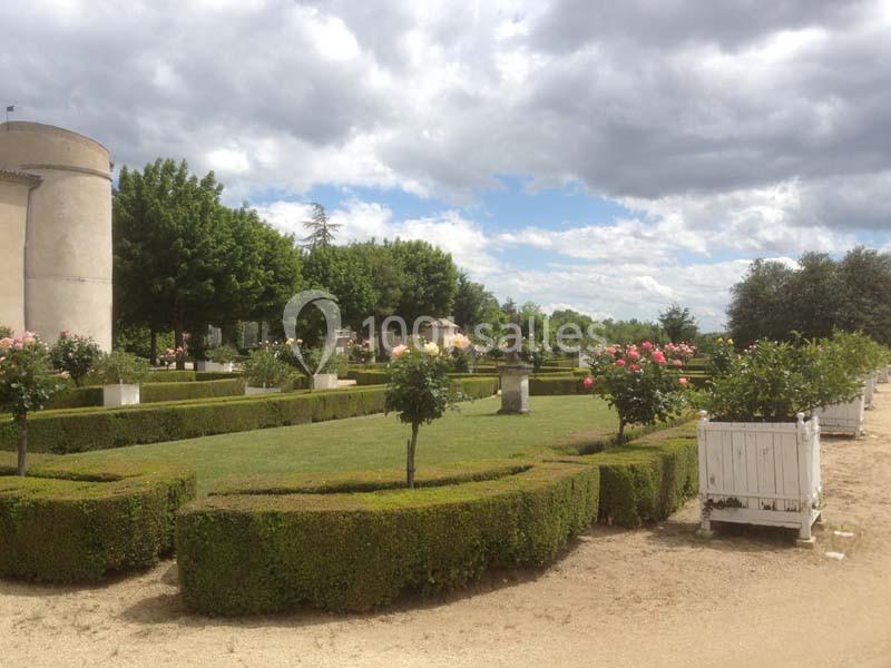 Jardin avec pelouse, haies taillées, rosiers en fleurs et allée sablonneuse sous un ciel nuageux.