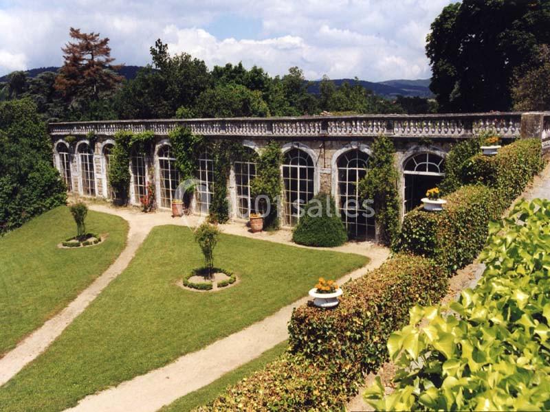 Serre ancienne avec de grandes fenêtres arquées entourée de jardins verdoyants et chemins gravillonnés.