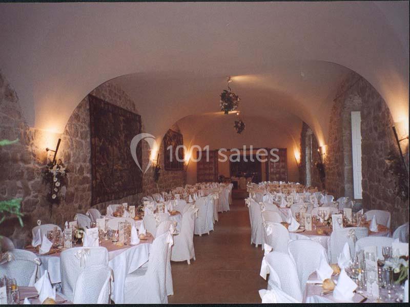 Salle voûtée en pierre avec tables rondes dressées pour un événement, décorée de nappes blanches et luminaires muraux.