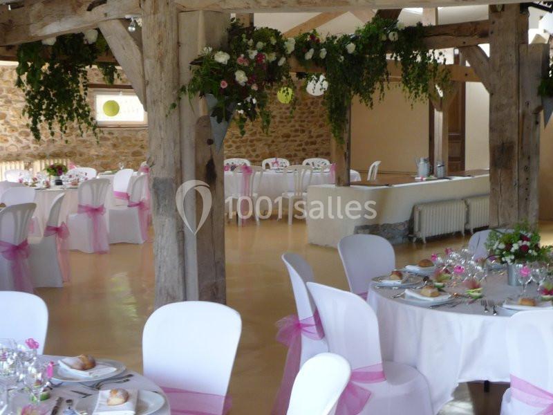 Salle de réception décorée avec des tables dressées, des chaises blanches ornées de nœuds roses et des guirlandes végétales.