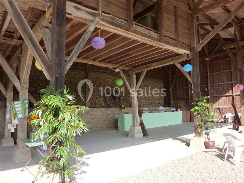 Intérieur d'une grange aménagée avec des tables décorées, des plantes et des lanternes colorées suspendues.