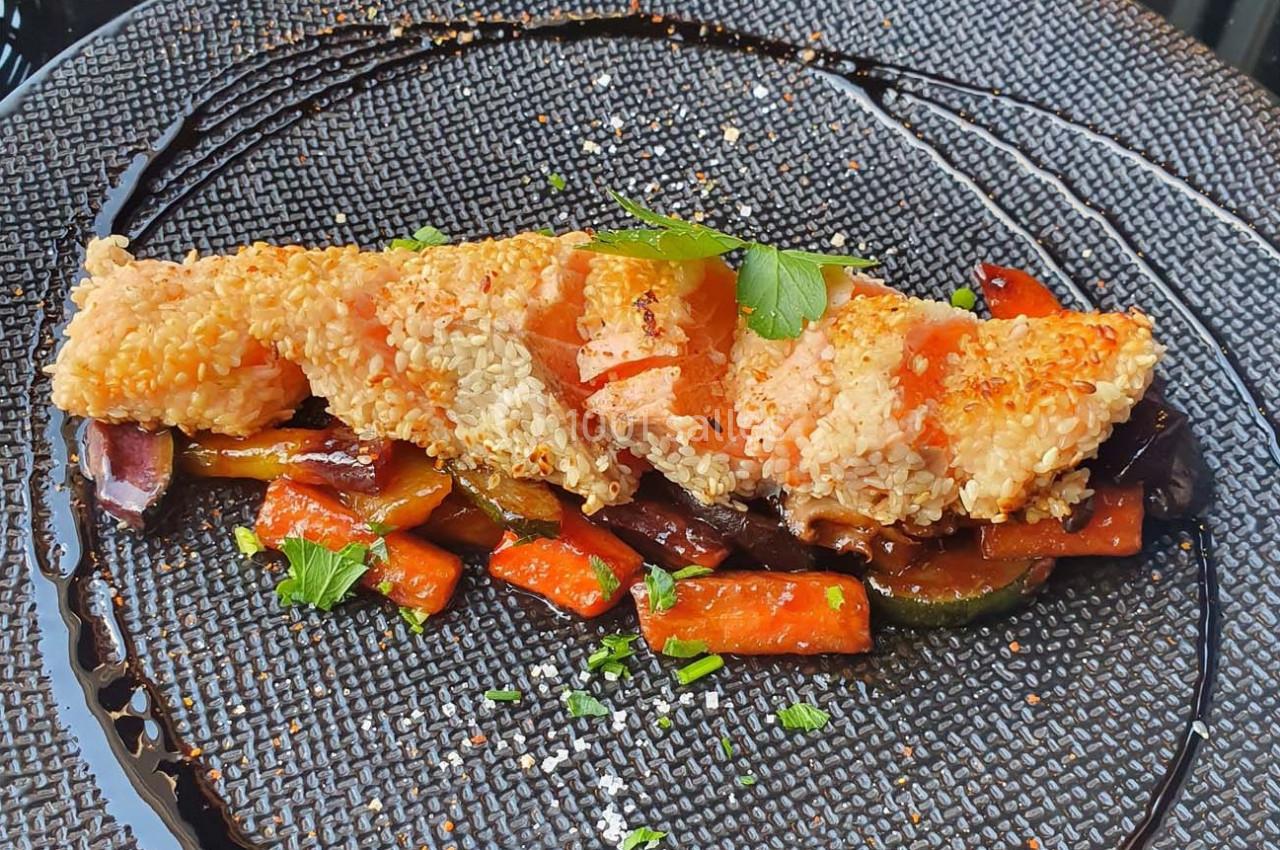 Filet de poisson pané au sésame servi sur un lit de légumes grillés, décoré de persil et d'un filet de sauce balsamique.