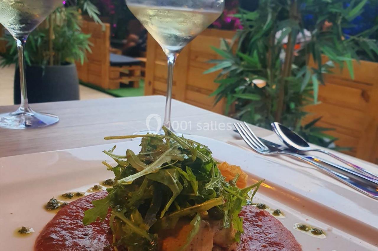 Assiette de viande garnie de roquette et sauce rouge, servie sur une table avec des verres de vin en arrière-plan.