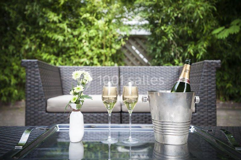 Seau à champagne, deux verres remplis et un vase de fleurs sur une table en extérieur, avec canapé et végétation en arrière…