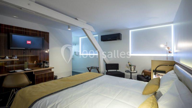 Chambre moderne avec lit double, bureau, télévision murale, fauteuils, table basse et lumière naturelle filtrée.