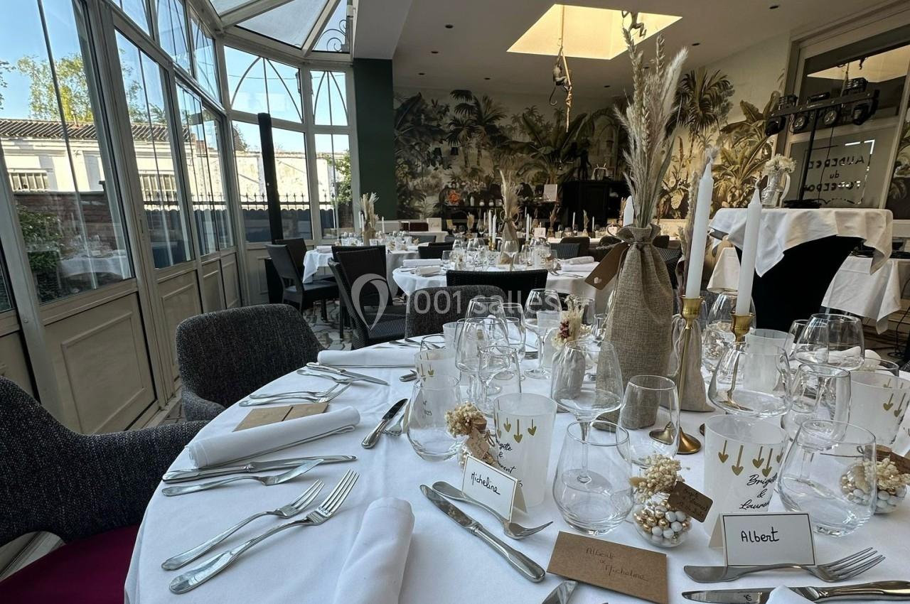 Salle de réception élégante avec tables rondes dressées, décorations florales et lumière naturelle provenant de grandes…