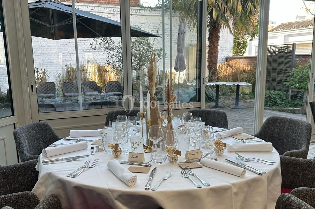 Table élégamment dressée avec nappes blanches, verres et bougies, dans une salle lumineuse donnant sur un jardin.