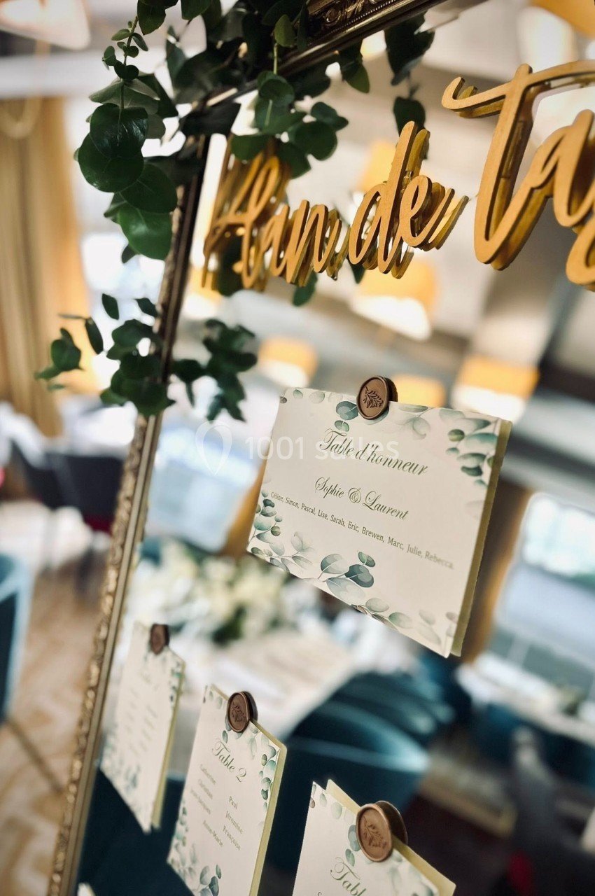 Plan de table décoratif avec cartes nominatives fixées sur un miroir entouré de feuillage, dans une salle élégante.