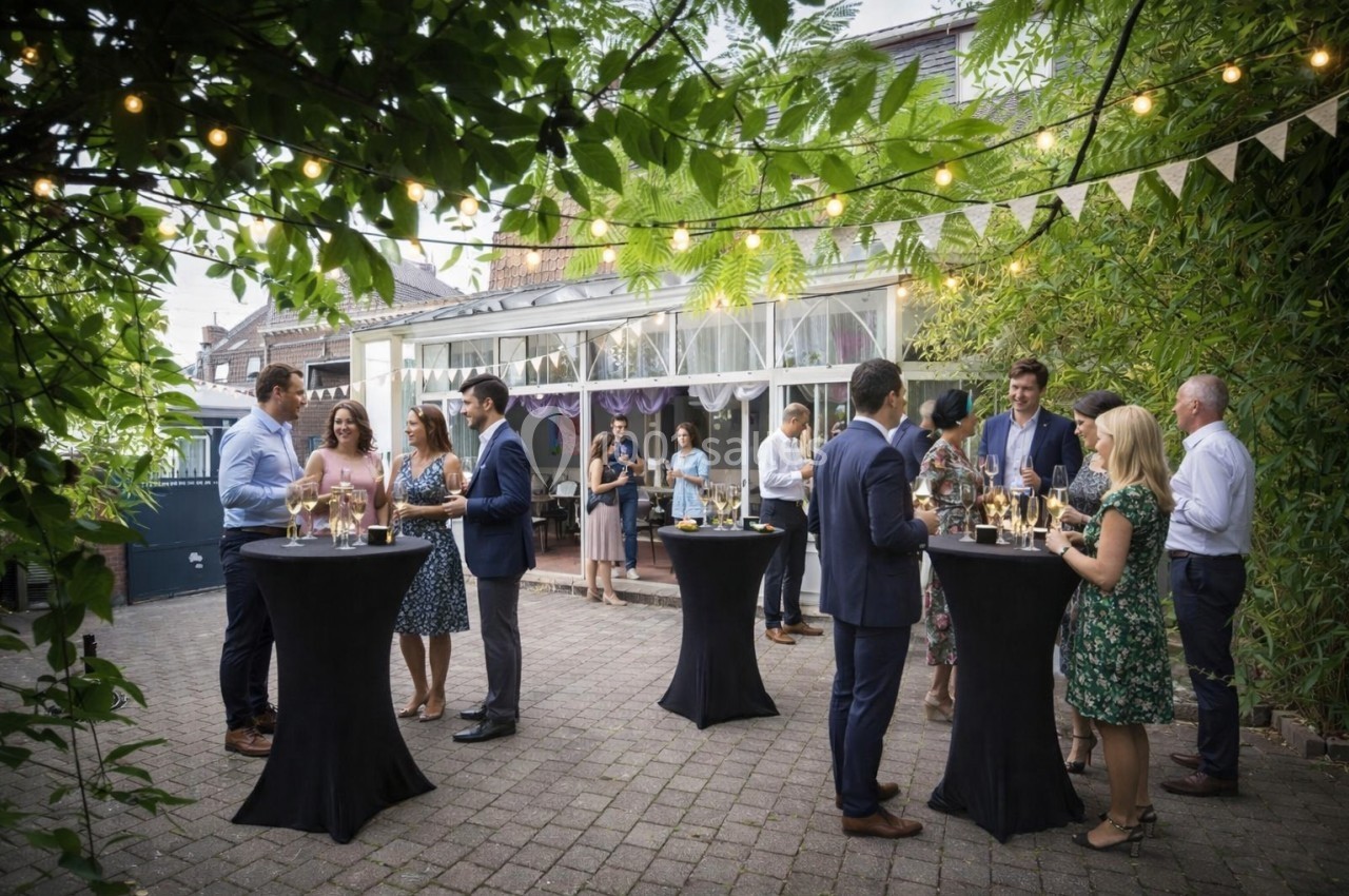 Des personnes discutent en petits groupes autour de tables hautes lors d'un événement en extérieur sous des guirlandes…