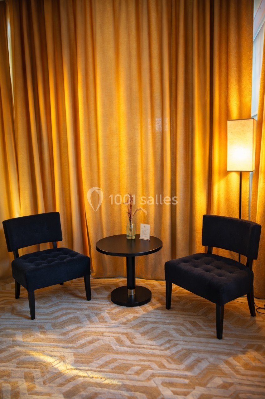 Deux fauteuils noirs entourent une table ronde avec un vase, devant des rideaux dorés et une lampe allumée.